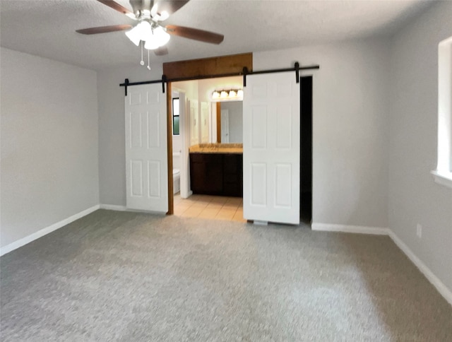 unfurnished bedroom featuring a barn door, ceiling fan, light colored carpet, and ensuite bath