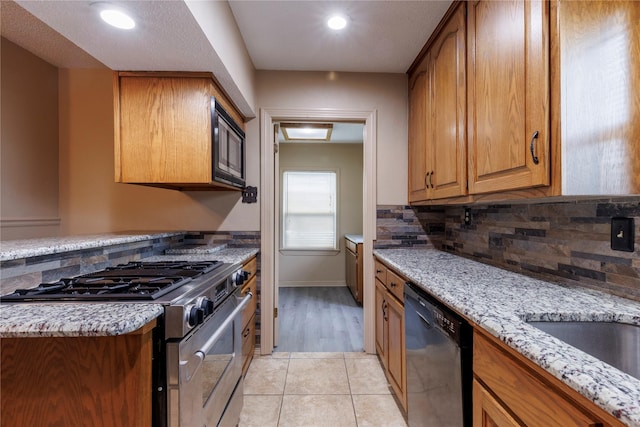 kitchen with backsplash, brown cabinetry, light tile patterned flooring, light stone countertops, and black appliances