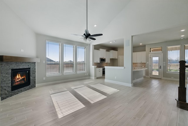 unfurnished living room featuring high vaulted ceiling, a stone fireplace, light wood-style flooring, a ceiling fan, and baseboards