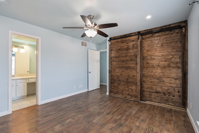 unfurnished bedroom with ensuite bathroom, ceiling fan, wood-type flooring, a barn door, and a closet