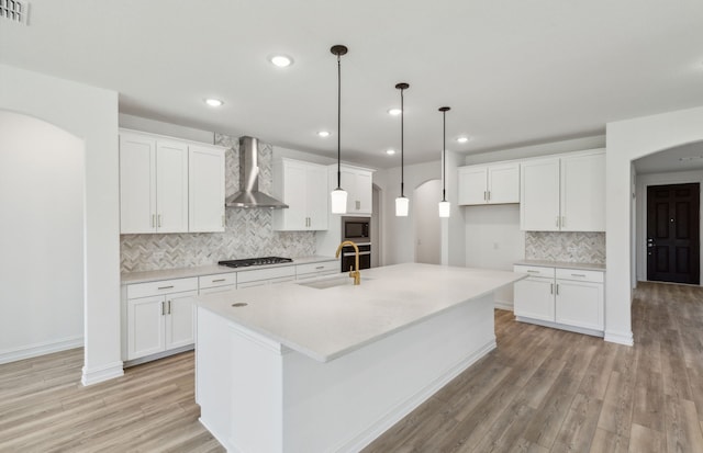 kitchen with wall chimney exhaust hood, white cabinets, light hardwood / wood-style floors, and a kitchen island with sink