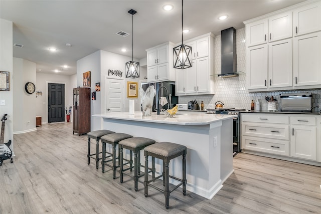 kitchen featuring wall chimney range hood, light hardwood / wood-style flooring, a kitchen island with sink, stainless steel gas stove, and pendant lighting