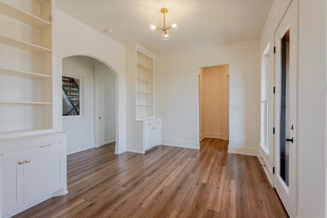unfurnished dining area featuring built in features, arched walkways, an inviting chandelier, and wood finished floors