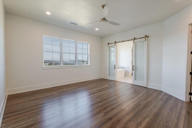 unfurnished bedroom featuring dark hardwood / wood-style flooring, a barn door, connected bathroom, and ceiling fan