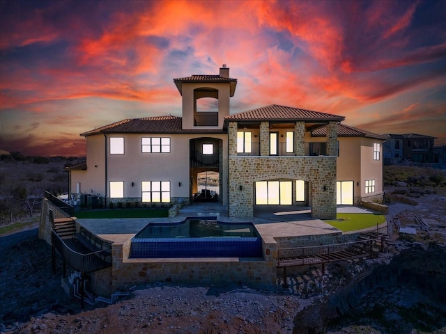 back of property at dusk featuring a balcony, a patio, stucco siding, a chimney, and a tile roof