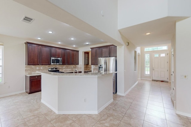 kitchen featuring light tile patterned flooring, a center island with sink, stainless steel appliances, light stone countertops, and decorative backsplash