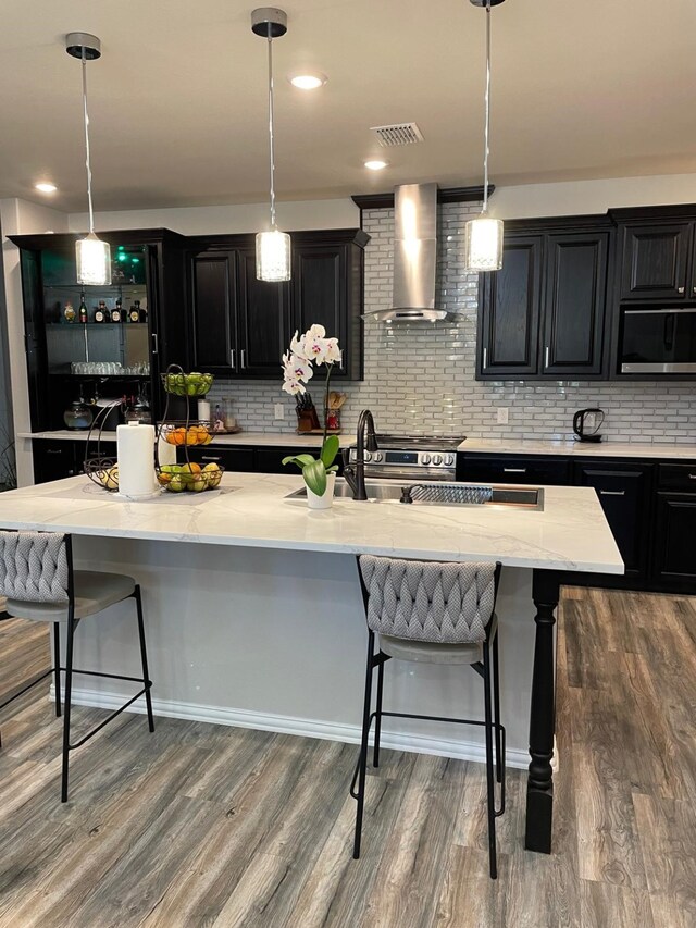 kitchen with a kitchen bar, appliances with stainless steel finishes, wall chimney range hood, decorative light fixtures, and an island with sink