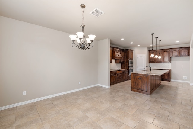 kitchen featuring an inviting chandelier, a center island with sink, light stone countertops, tasteful backsplash, and decorative light fixtures