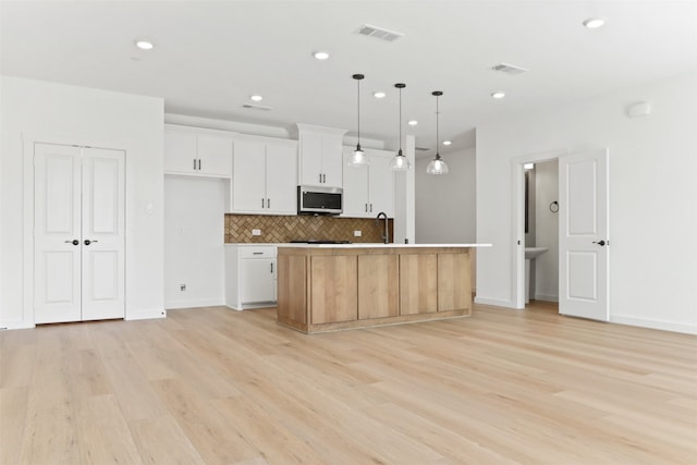 kitchen featuring light wood-type flooring, an island with sink, hanging light fixtures, white cabinetry, and decorative backsplash