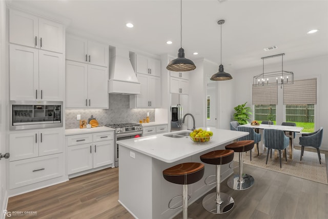 kitchen with appliances with stainless steel finishes, premium range hood, white cabinets, hanging light fixtures, and an island with sink