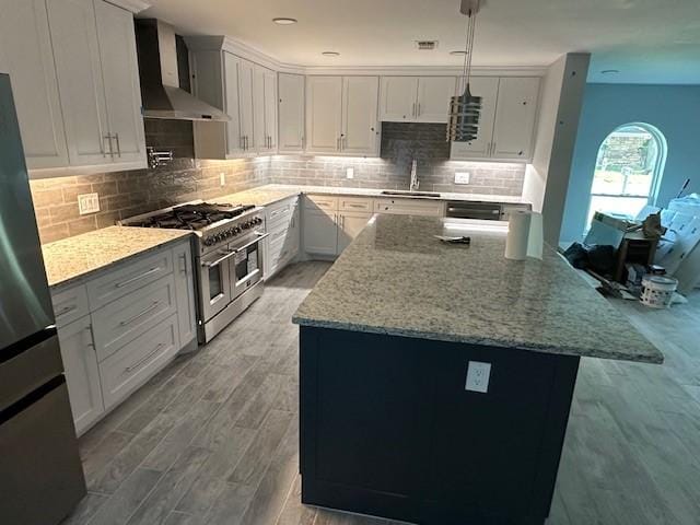 kitchen featuring wall chimney range hood, white cabinetry, hanging light fixtures, light stone countertops, and appliances with stainless steel finishes