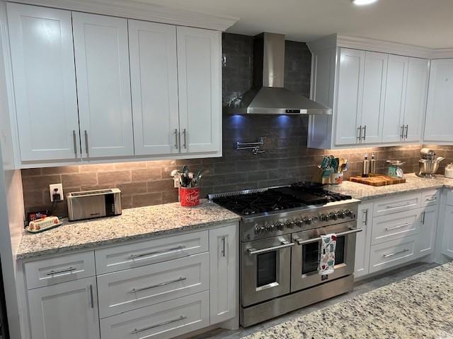 kitchen with tasteful backsplash, wall chimney exhaust hood, range with two ovens, and white cabinetry