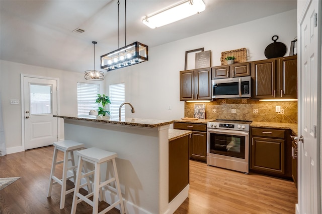 kitchen with light hardwood / wood-style flooring, appliances with stainless steel finishes, a kitchen island with sink, a kitchen bar, and decorative light fixtures