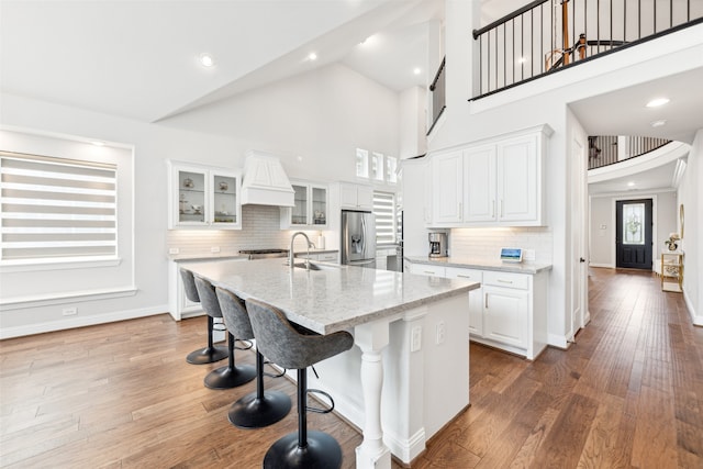 kitchen featuring white cabinetry, light stone counters, high vaulted ceiling, stainless steel refrigerator with ice dispenser, and an island with sink