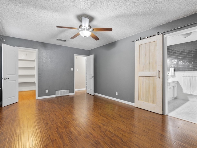 unfurnished bedroom featuring ensuite bath, dark wood-type flooring, a textured ceiling, a walk in closet, and a barn door