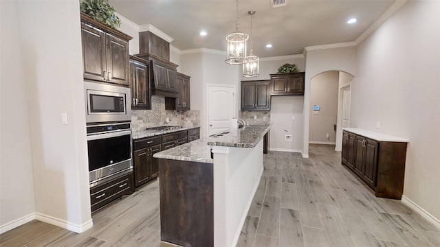 kitchen with hanging light fixtures, ornamental molding, stainless steel appliances, a kitchen island with sink, and backsplash