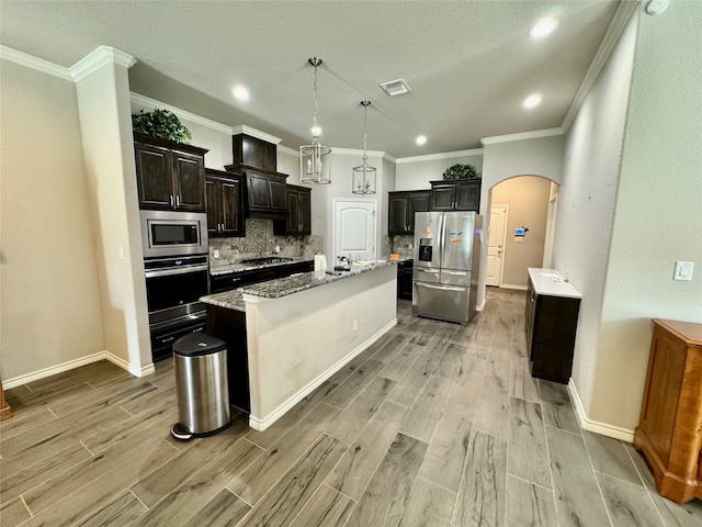 kitchen with ornamental molding, appliances with stainless steel finishes, a kitchen island with sink, decorative light fixtures, and tasteful backsplash