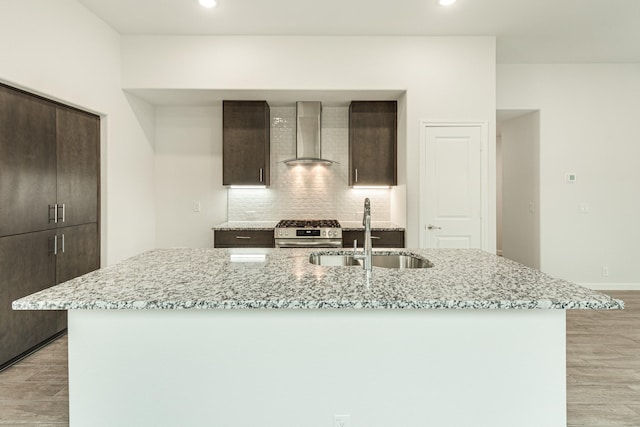 kitchen featuring stainless steel gas range oven, light stone counters, wall chimney range hood, and an island with sink