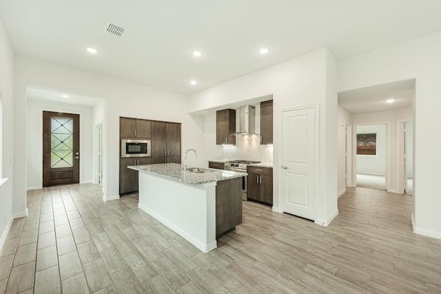 kitchen featuring an island with sink, light hardwood / wood-style floors, wall chimney range hood, and appliances with stainless steel finishes