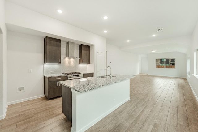 kitchen featuring wall chimney exhaust hood, a kitchen island with sink, sink, light hardwood / wood-style floors, and stainless steel range with gas cooktop