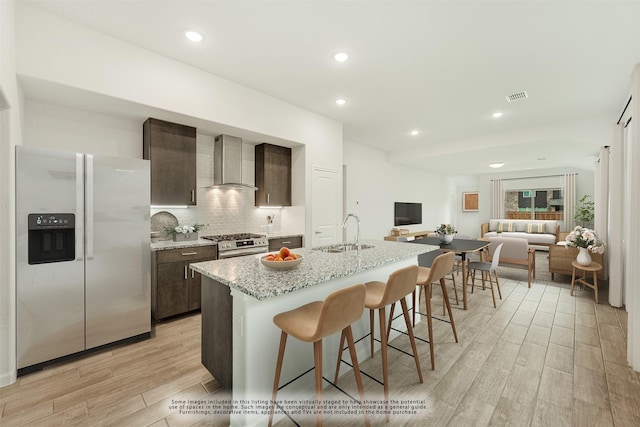 kitchen featuring sink, wall chimney exhaust hood, an island with sink, dark brown cabinetry, and stainless steel appliances