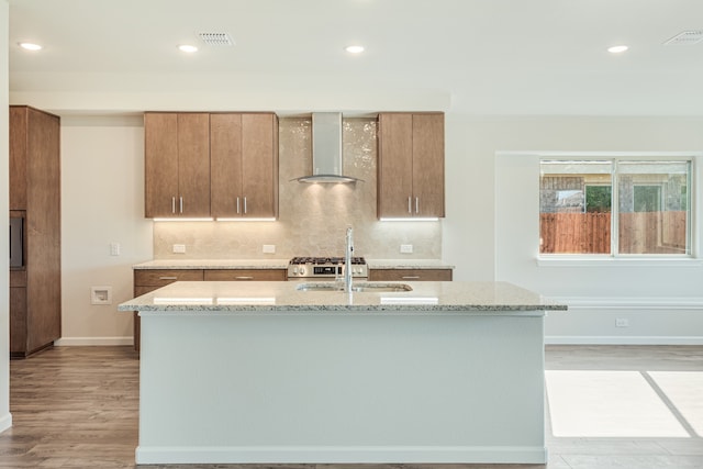 kitchen with light wood-type flooring, a center island with sink, and wall chimney range hood