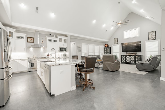 kitchen featuring an island with sink, appliances with stainless steel finishes, white cabinets, wall chimney exhaust hood, and ceiling fan