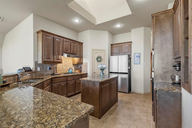 kitchen with dark stone countertops, a sink, stainless steel appliances, under cabinet range hood, and backsplash