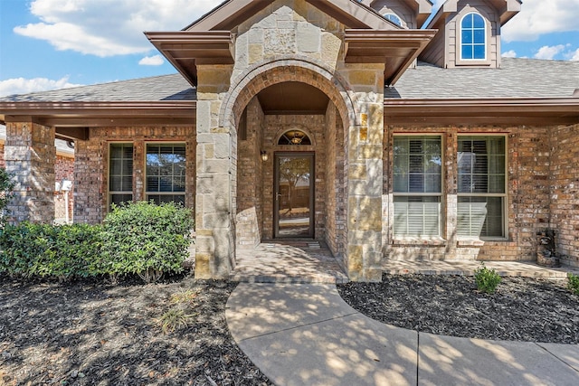 view of exterior entry with stone siding, a shingled roof, and brick siding