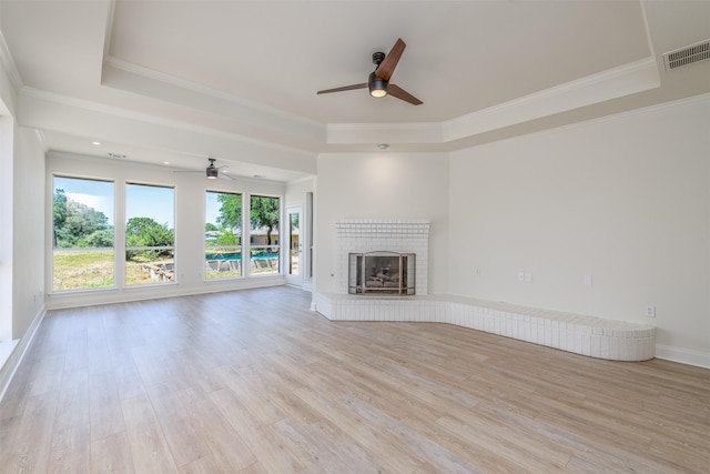 unfurnished living room featuring a brick fireplace, ornamental molding, a tray ceiling, ceiling fan, and light hardwood / wood-style floors