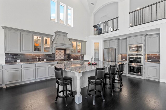 kitchen featuring decorative backsplash, premium range hood, a kitchen island with sink, high vaulted ceiling, and gray cabinets