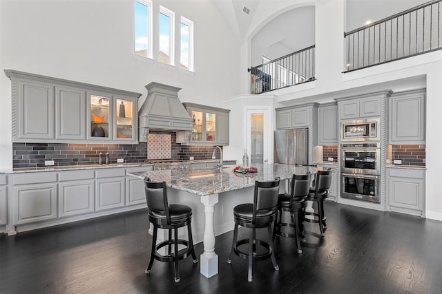 kitchen with gray cabinets, an island with sink, custom range hood, stainless steel appliances, and light stone countertops