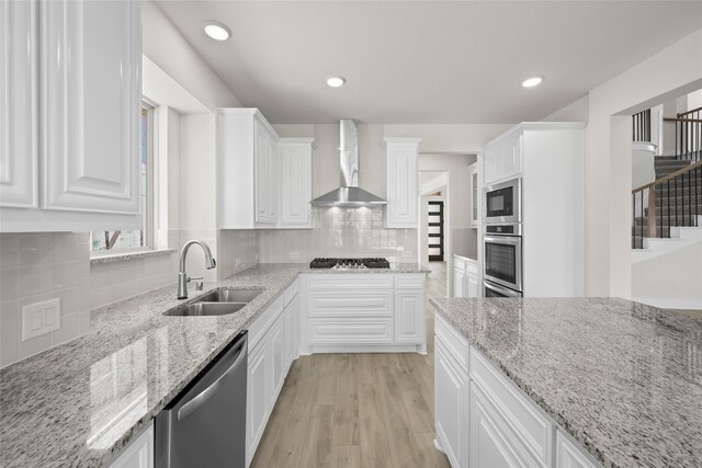 kitchen with light stone countertops, appliances with stainless steel finishes, wall chimney exhaust hood, sink, and white cabinets