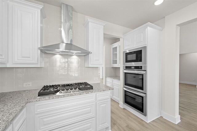 kitchen with white cabinets, wall chimney exhaust hood, light stone countertops, light hardwood / wood-style floors, and stainless steel appliances