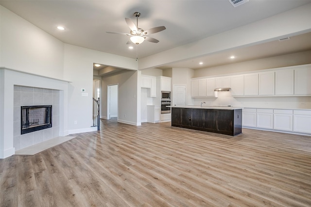 kitchen with a tile fireplace, a center island with sink, white cabinets, light wood-type flooring, and appliances with stainless steel finishes