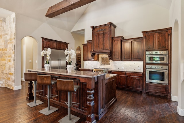 kitchen featuring beamed ceiling, an island with sink, appliances with stainless steel finishes, and tasteful backsplash