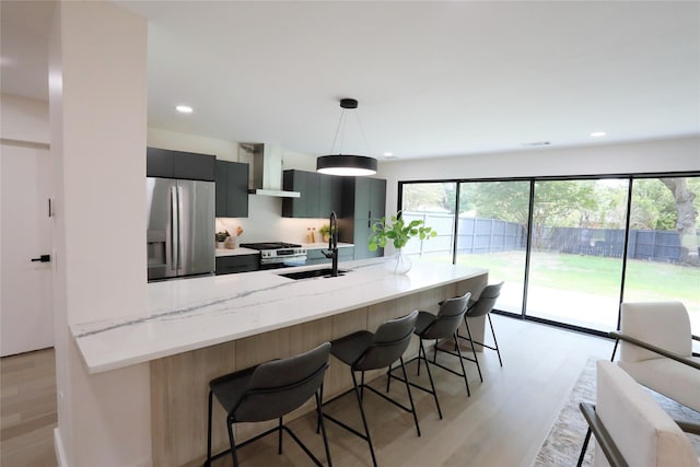 kitchen featuring wall chimney range hood, sink, hanging light fixtures, stainless steel appliances, and light stone counters