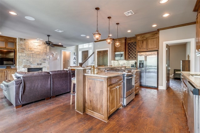 kitchen featuring appliances with stainless steel finishes, decorative light fixtures, light stone counters, a kitchen island with sink, and a stone fireplace