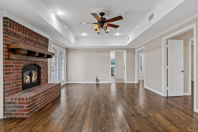 unfurnished living room featuring a brick fireplace, ceiling fan, a tray ceiling, crown molding, and dark wood-type flooring