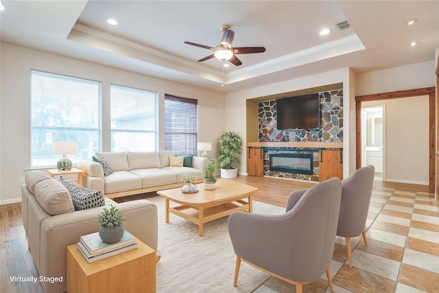 living room with light wood-type flooring, a stone fireplace, a tray ceiling, and ceiling fan