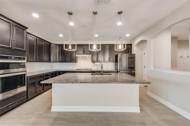 kitchen with sink, a kitchen island with sink, dark stone countertops, stainless steel appliances, and decorative light fixtures