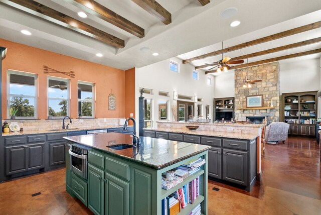 kitchen featuring a center island with sink, beamed ceiling, a stone fireplace, sink, and ceiling fan