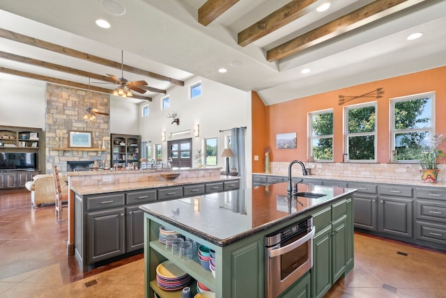 kitchen featuring beamed ceiling, a kitchen island with sink, a stone fireplace, stainless steel oven, and ceiling fan