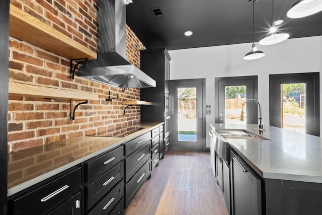 kitchen with black electric cooktop, a kitchen island with sink, sink, decorative light fixtures, and wall chimney range hood