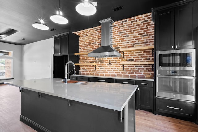 kitchen featuring pendant lighting, a center island with sink, stainless steel microwave, light hardwood / wood-style floors, and wall chimney exhaust hood