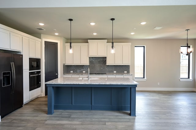 kitchen with white cabinets, hanging light fixtures, light stone counters, black appliances, and a center island with sink