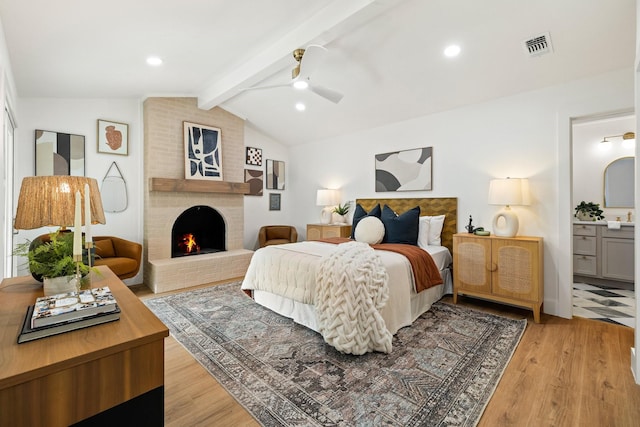 bedroom with ensuite bath, ceiling fan, a brick fireplace, lofted ceiling with beams, and light wood-type flooring