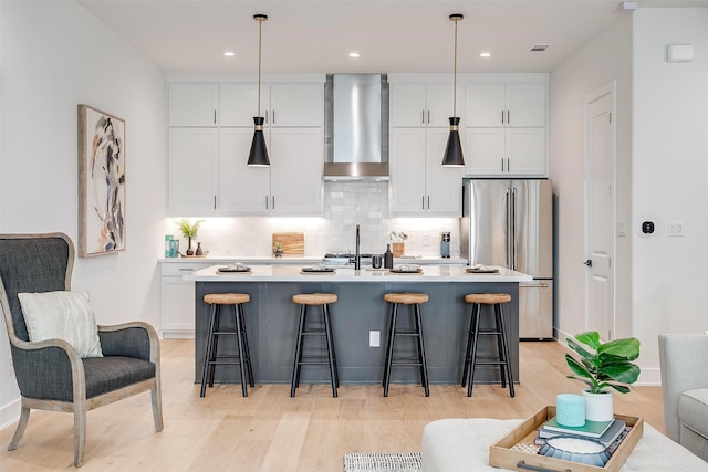 kitchen featuring a kitchen island with sink, wall chimney range hood, hanging light fixtures, white cabinetry, and stainless steel refrigerator