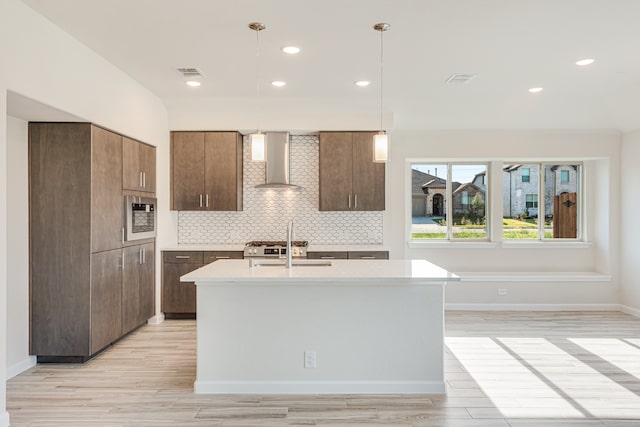 kitchen featuring backsplash, light hardwood / wood-style flooring, a center island with sink, pendant lighting, and wall chimney range hood