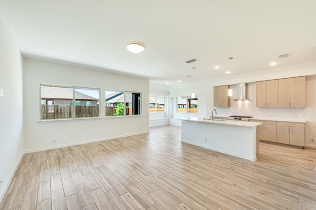 kitchen with decorative light fixtures, wall chimney exhaust hood, light brown cabinetry, an island with sink, and tasteful backsplash